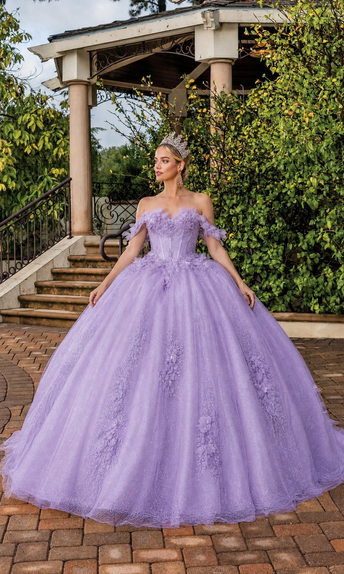 A woman wearing the Lilac Quinceanera Dress 1883 by Dancing Queen, featuring a sweetheart neckline and paired with a tiara, stands on a brick path in front of a gazebo surrounded by lush greenery.